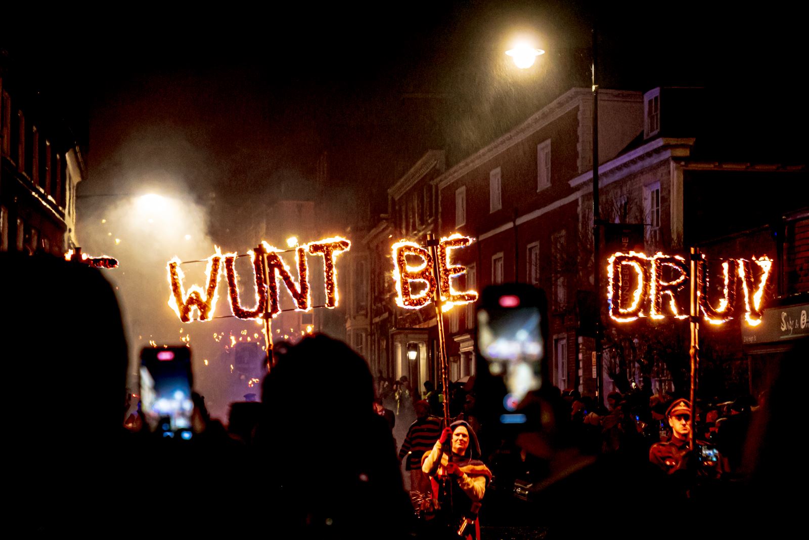 Torchlit bonfire procession in Sussex, with participants carrying illuminated letters spelling “Wunt Be Druv”, the traditional county motto meaning “We will not be driven”, as crowds gather along the street at night.